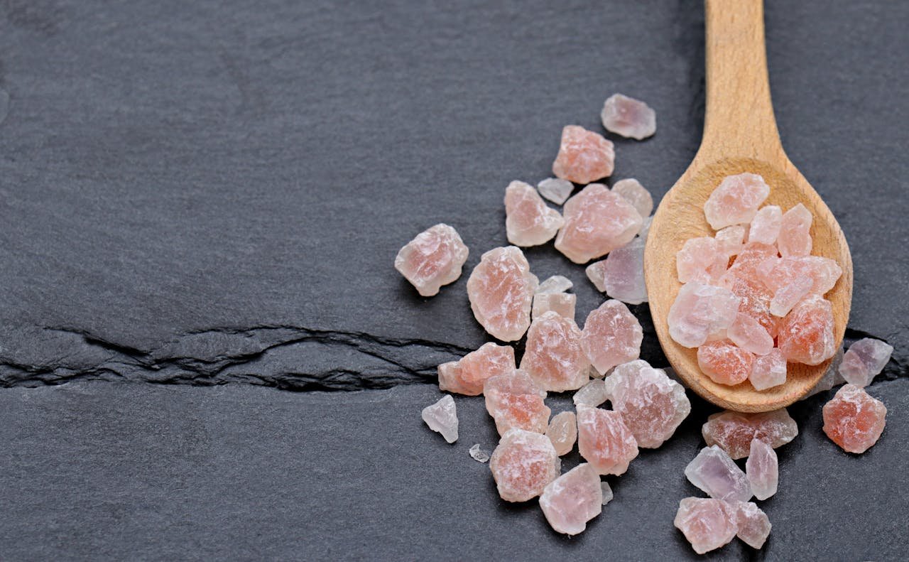 A wooden spoon with Himalayan pink salt crystals on a dark slate surface.